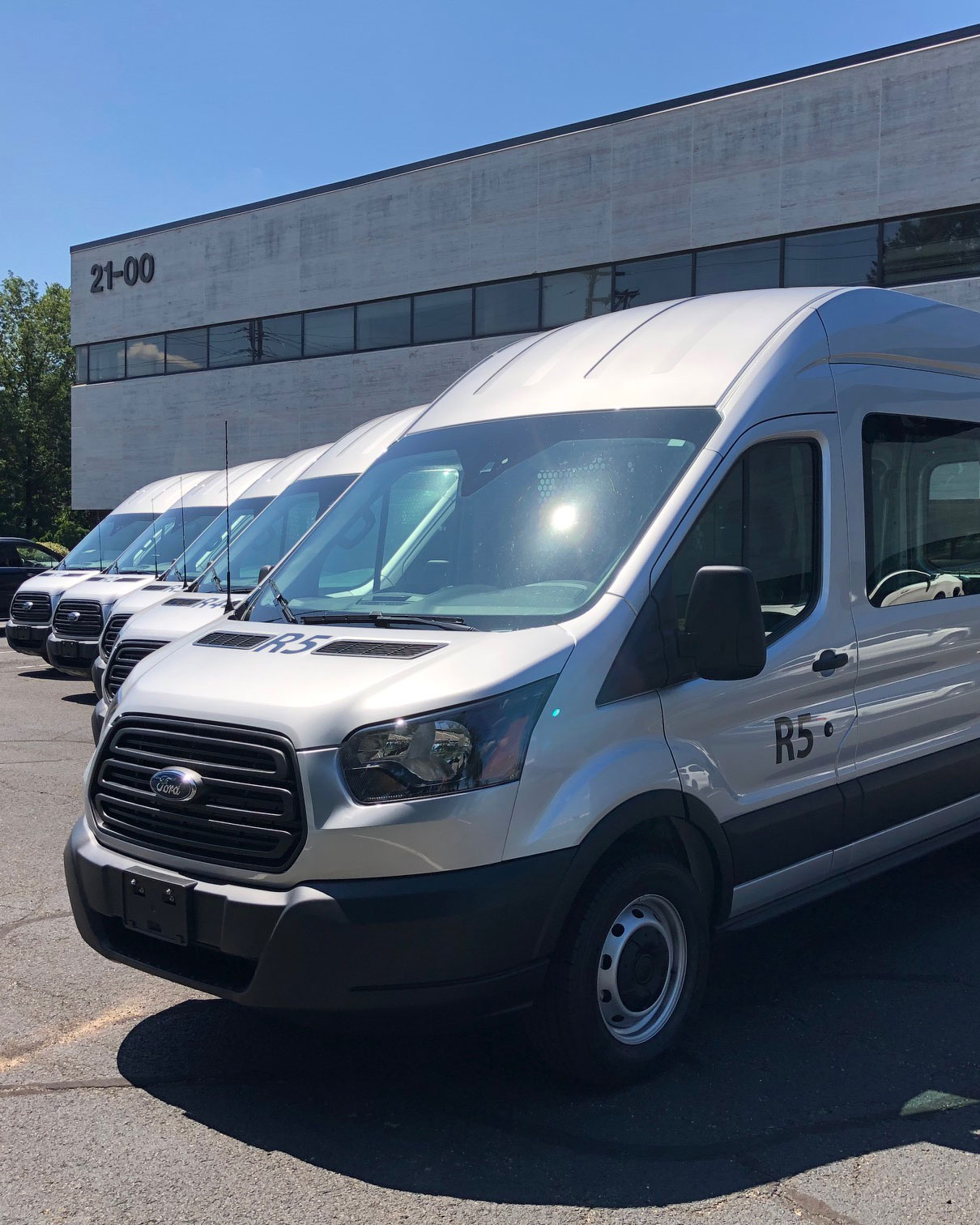 A row of parked white Ford vans is lined up in front of a modern office building with large windows. The vans have identification numbers such as "R5" on their front. The sky is clear and blue.